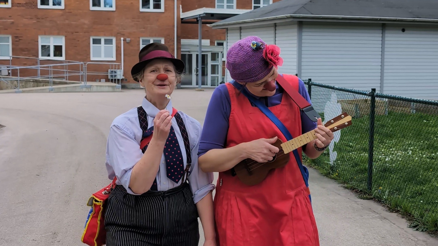 Foto på två sjukhusclowner utanför palliativa avdelningen i Lund. Den ena spelar ukulele och den andra håller i en blomma.